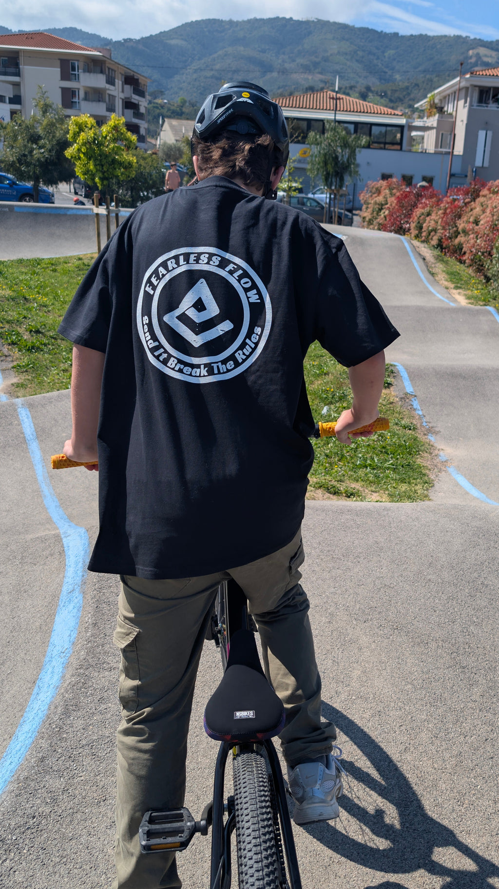 Rider VTT portant un tee-shirt streetwear Fearless Flow noir avec logo dos Send It Break The Rules sur un pump track en extérieur