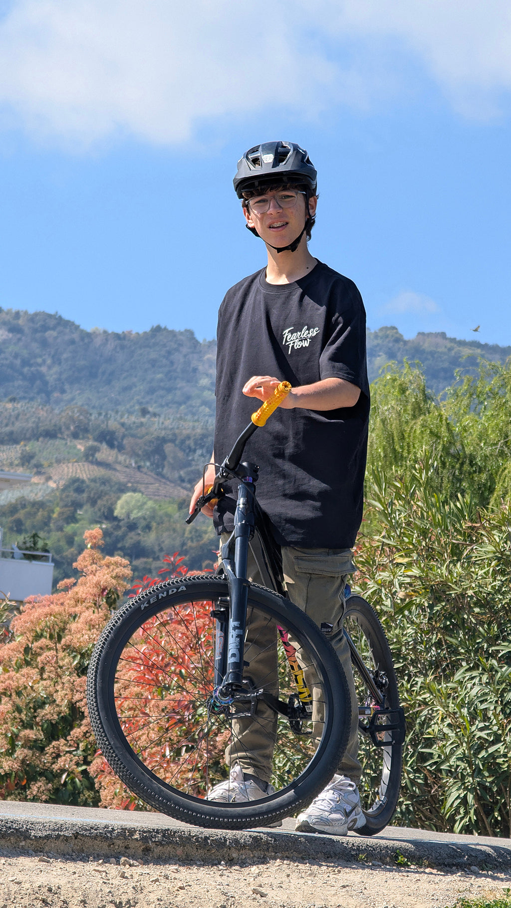 Jeune rider VTT portant un tee-shirt streetwear Fearless Flow noir avec logo poitrine, debout sur un pump track avec son vélo devant un paysage de montagnes