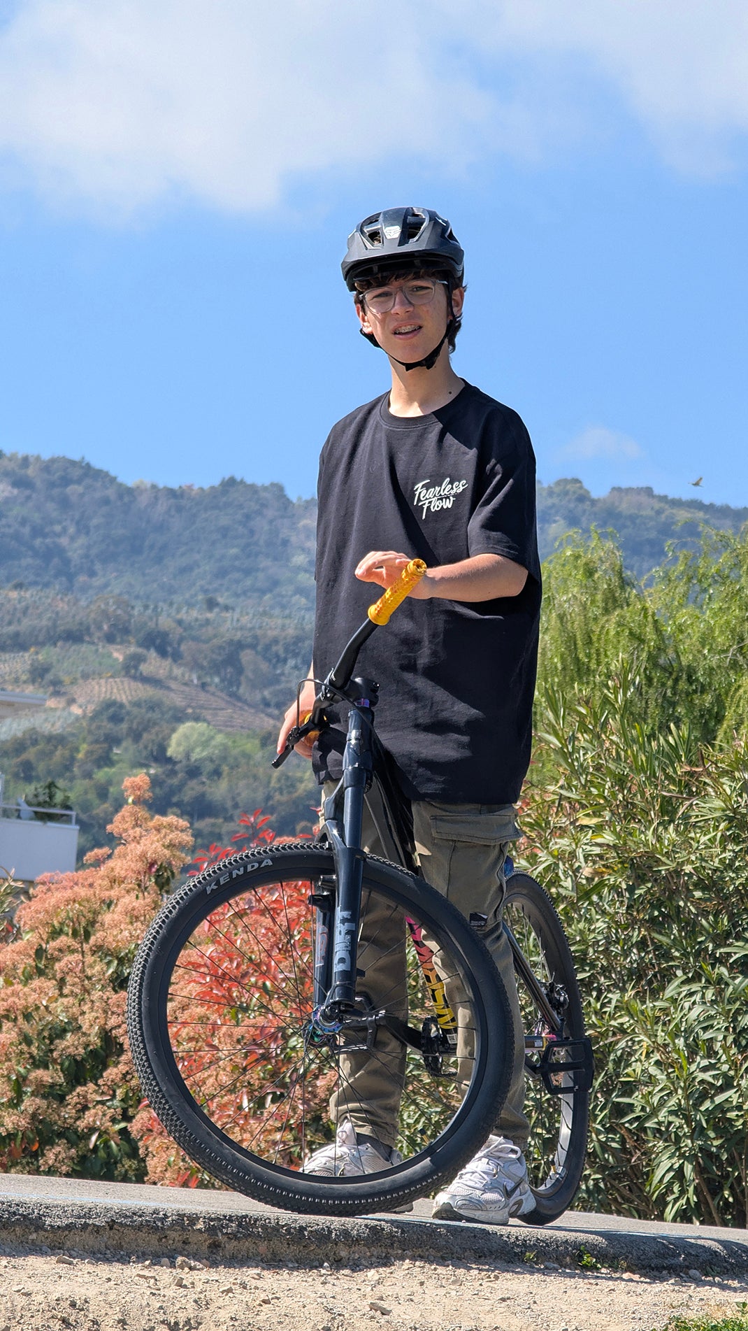 Jeune rider VTT portant un tee-shirt streetwear Fearless Flow noir avec logo poitrine, debout sur un pump track avec son vélo devant un paysage de montagnes